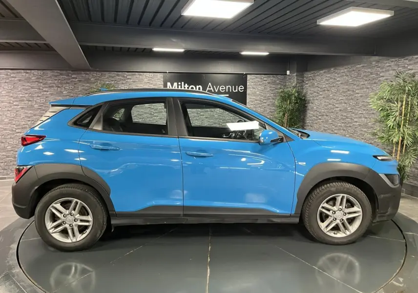 Hyundai Kona bleu en profil côté gauche, avec protections noires et jantes alliage 16 pouces visibles en intérieur showroom.