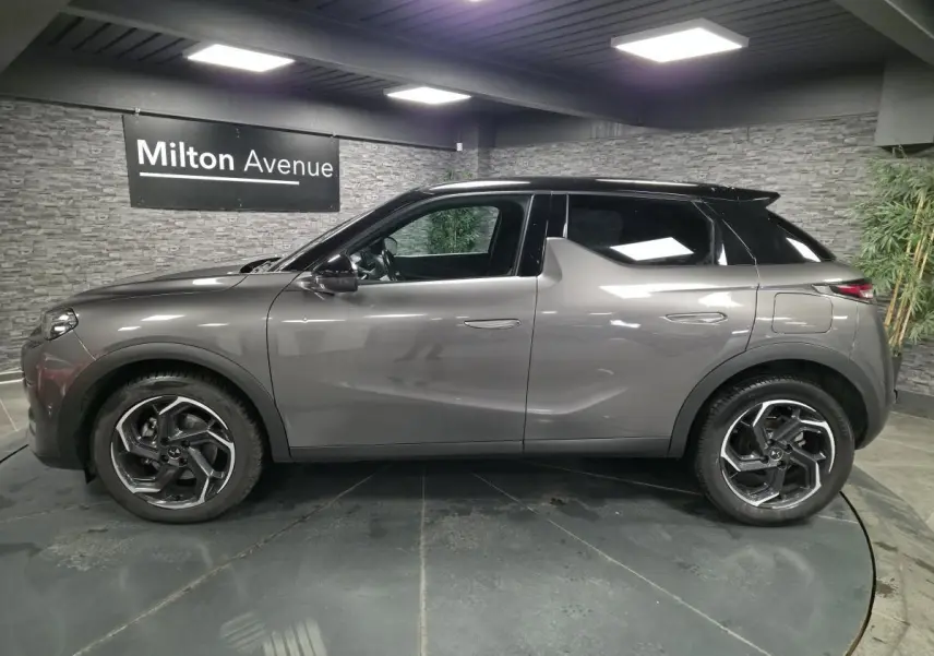 Vue de profil côté gauche du DS3 Crossback gris métallisé avec toit noir et jantes alliage 18 pouces dans un showroom.