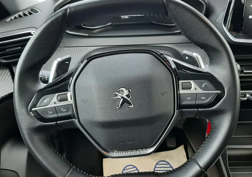 Vue rapprochée du volant noir en cuir du Peugeot 2008 blanc, avec commandes intégrées et tableau de bord en arrière-plan.
