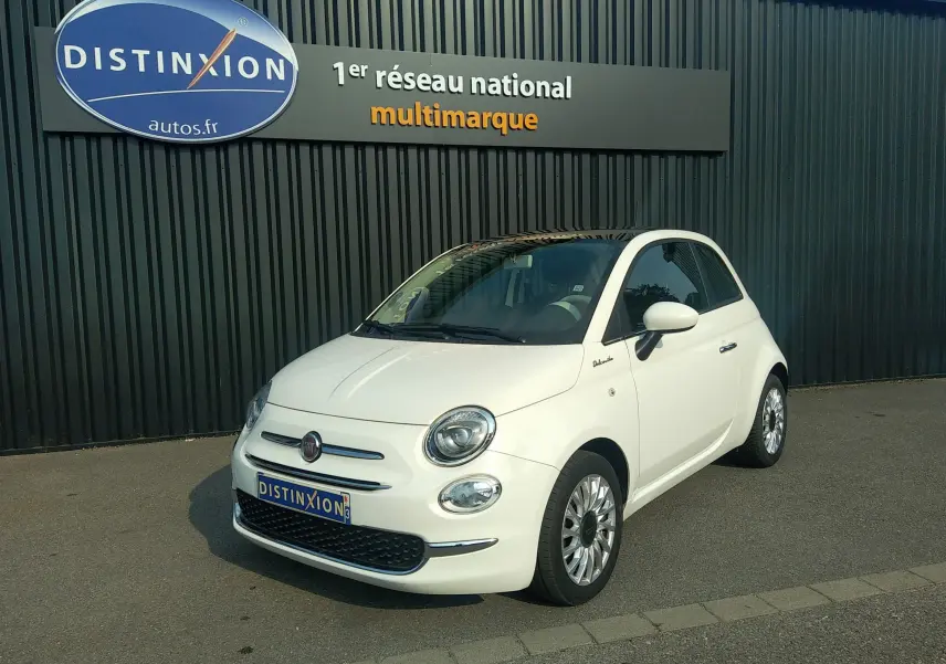 Fiat 500 blanche vue en 3/4 avant droit, avec toit noir et jantes alliage, stationnée devant un mur sombre.