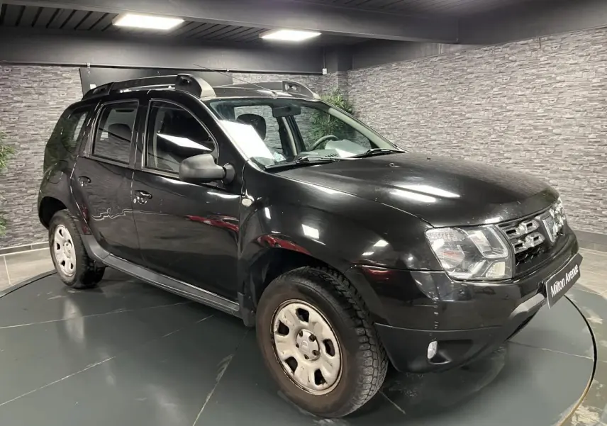 Dacia Duster noir vue 3/4 avant droit en intérieur avec barres de toit et jantes acier 16 pouces.