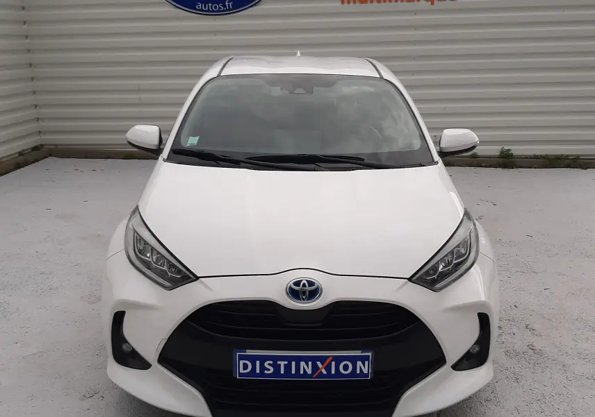 Vue frontale d'une Toyota Yaris blanche 2023 avec calandre noire et phares LED, stationnée devant un mur gris.