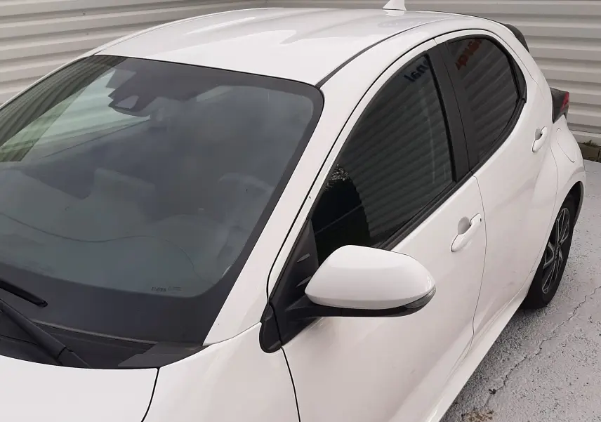Toyota Yaris blanche vue 3/4 avant droit, avec vitres teintées et rétroviseur blanc sur fond gris.
