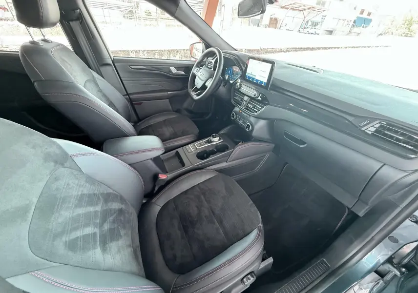 Vue intérieure côté passager avant du Ford Kuga bleu azur, sièges noirs avec surpiqûres rouges et tableau de bord moderne.