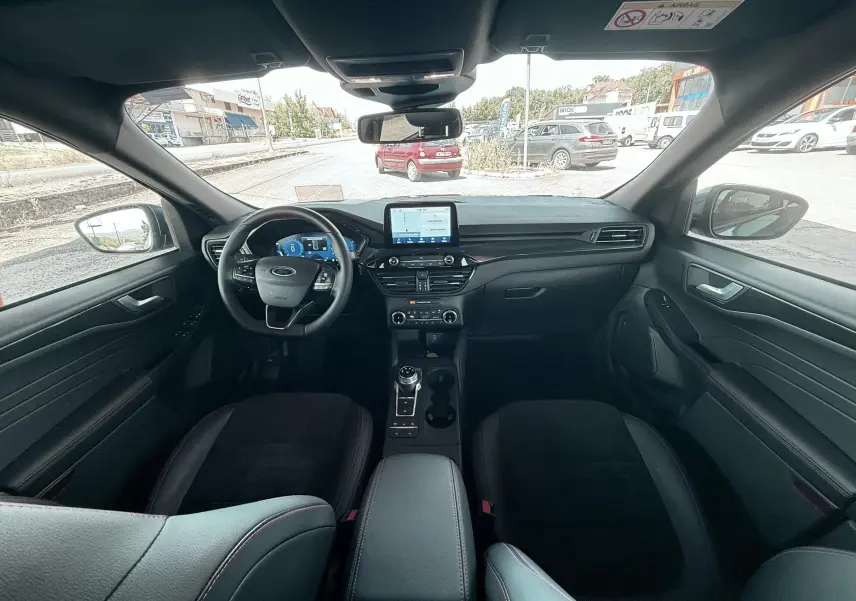 Vue intérieure avant du Ford Kuga 2023 avec tableau de bord noir, écran tactile central et volant ST-Line à surpiqûres rouges.