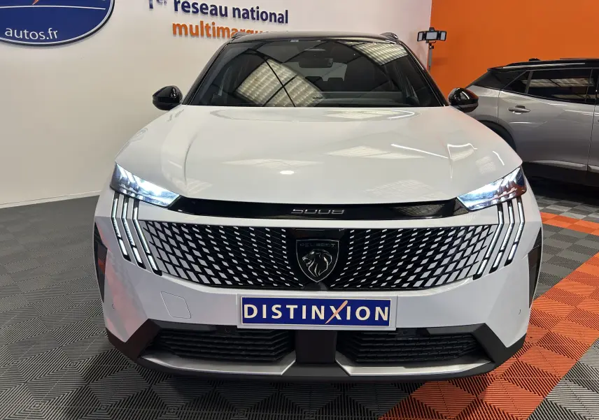 Vue de face du Peugeot 5008 Hybrid blanc 2025 avec calandre distinctive et phares LED allumés en intérieur showroom.