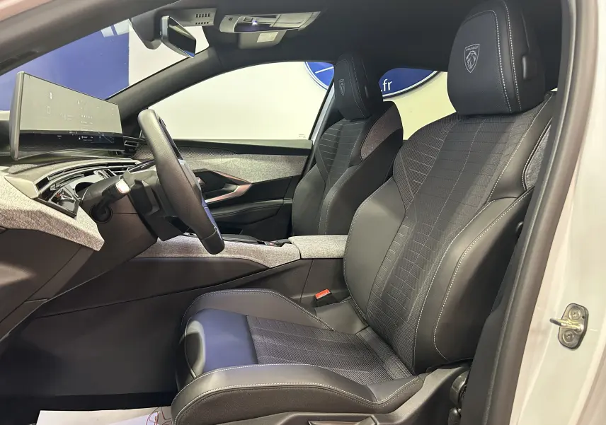 Vue intérieure côté conducteur du Peugeot 5008 Hybrid blanc 2025, sièges noirs avec logo Peugeot brodé sur appuie-tête.