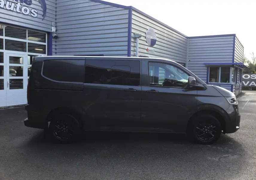 Profil latéral droit d'un Volkswagen Transporter ProCab gris anthracite avec jantes alu noires devant un bâtiment industriel.