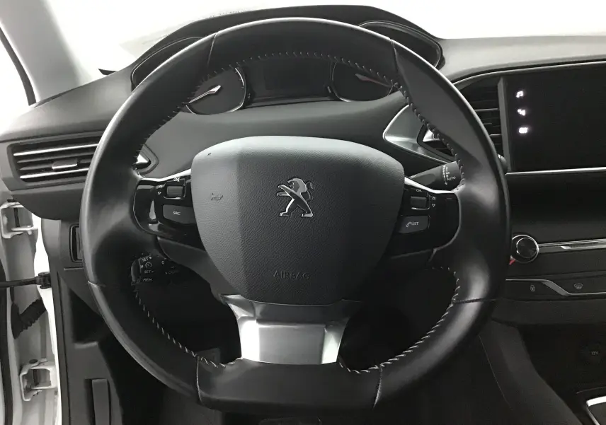 Vue rapprochée du volant cuir noir avec logo Peugeot au centre dans l'habitacle d'une 308 SW blanche de 2018.