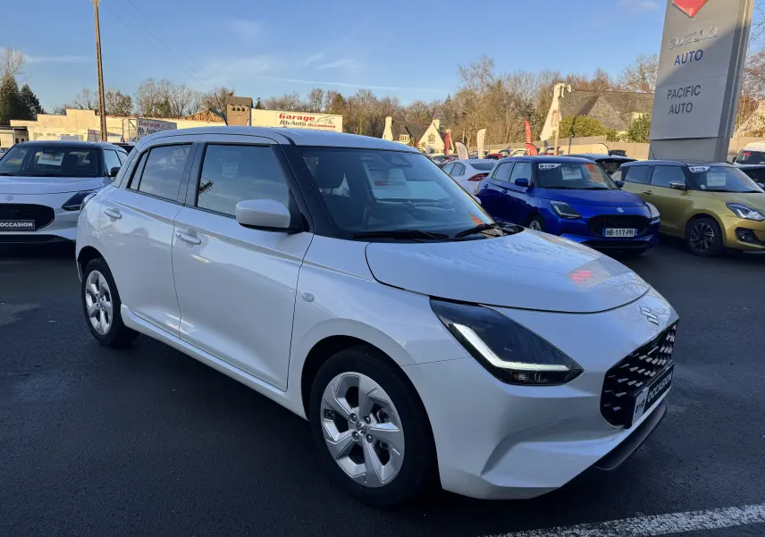 Vue 3/4 avant d'une Suzuki Swift blanche 2025 hybride essence, garée en extérieur sur un parking de concession.