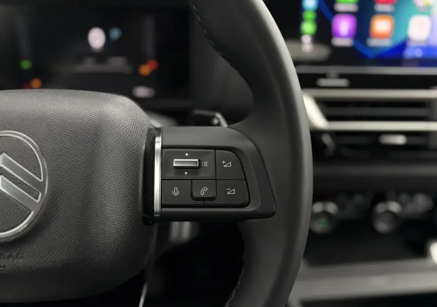 Gros plan sur le volant cuir noir de la Citroën C4 Hybrid 2025 avec commandes vocales et écran tactile flou en arrière-plan.