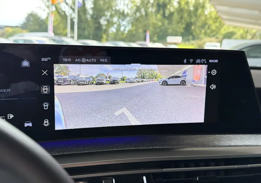 Vue écran caméra de recul affichant un parking extérieur avec plusieurs véhicules, prise depuis l'intérieur du Peugeot 1007 gris Artense.