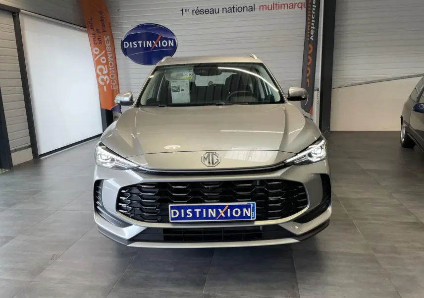 Vue frontale du MG ZS 1.5 Hybrid+ gris clair avec calandre noire et phares LED allumés en showroom.