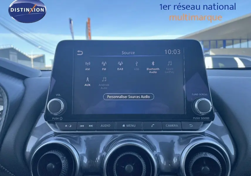 Écran tactile central du Nissan Juke gris squale métal 2024 affichant les sources audio, avec commandes et aérateurs visibles.