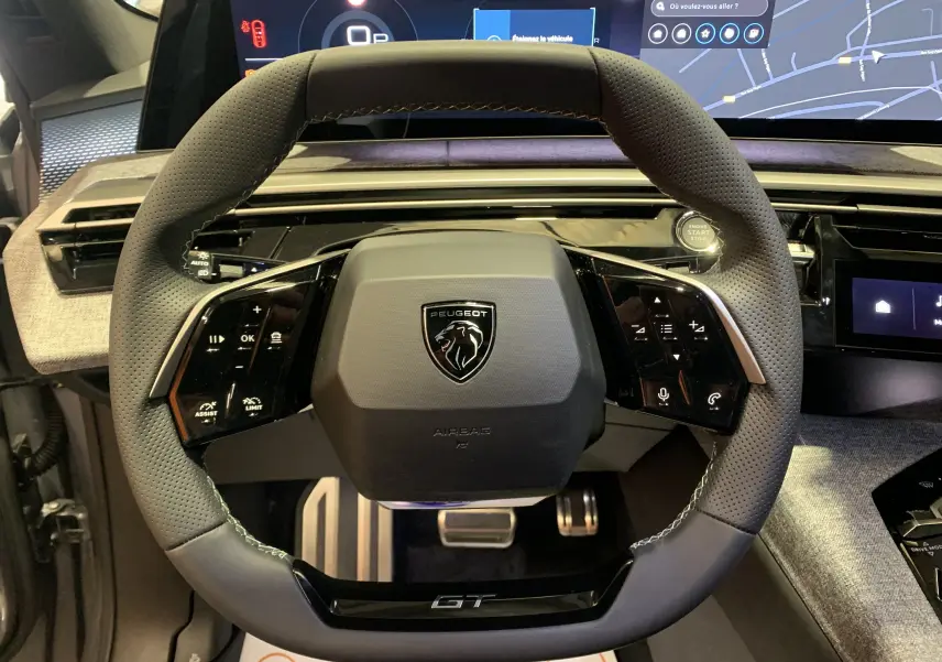 Vue rapprochée du volant GT en cuir perforé du Peugeot 3008 III Hybrid 136, avec tableau de bord digital en arrière-plan.