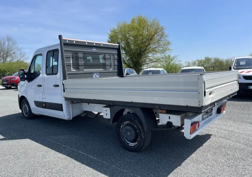 Renault Master blanc en 3/4 arrière droit, plateau ridelles aluminium et cabine double visible en extérieur sous ciel bleu.
