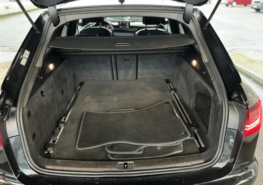 Vue arrière du coffre ouvert d'une Audi A6 Avant Allroad noire, avec tapis de sol noirs et intérieur spacieux.