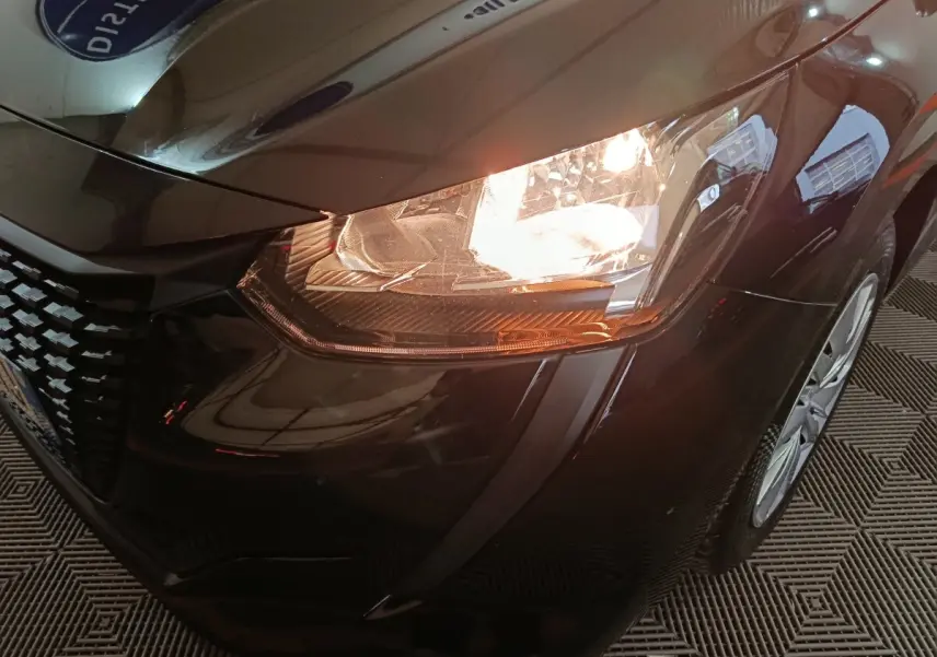Gros plan sur le phare avant droit allumé d'une Peugeot 208 noire, mettant en valeur la calandre et la jante avant.