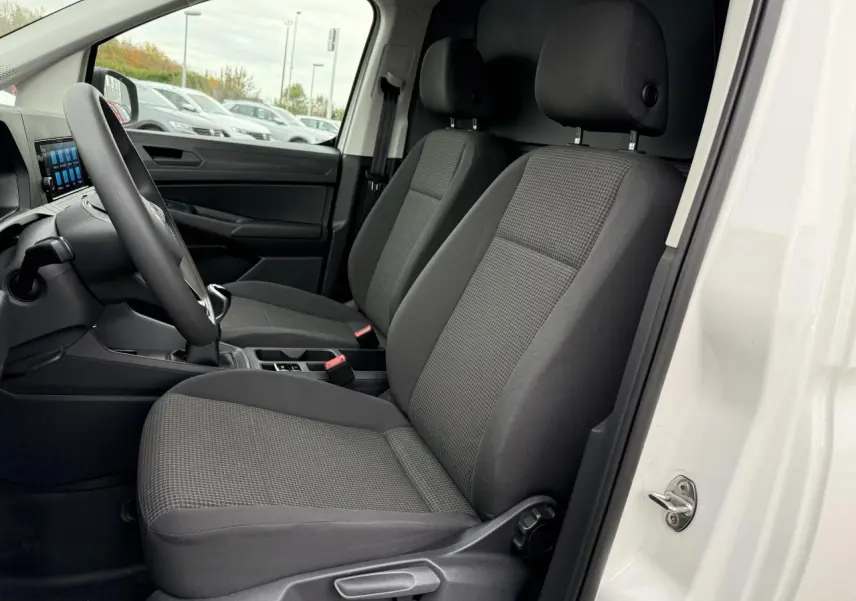 Vue intérieure côté conducteur du Volkswagen Caddy Maxi Cargo blanc candy, montrant les sièges avant en tissu gris et le volant multifonctions.