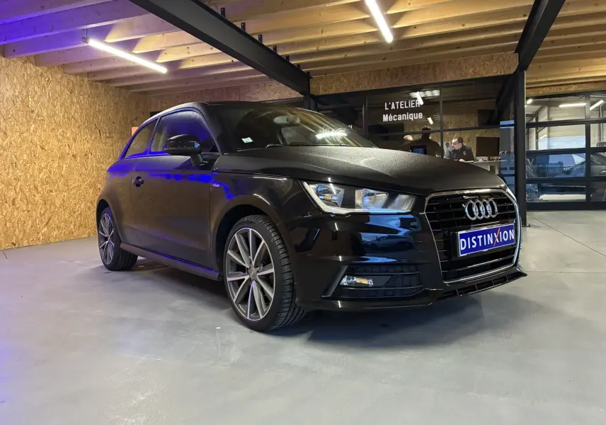 Audi A1 noir en vue 3/4 avant droit, avec jantes alliage et calandre distinctive S-Line dans un atelier.