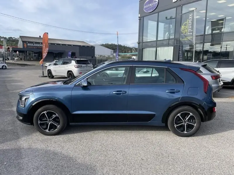 Vue de profil côté gauche d'un KIA NIRO 2024 bleu métal, hybride essence avec jantes noires et détails noirs sur les bas de caisse.