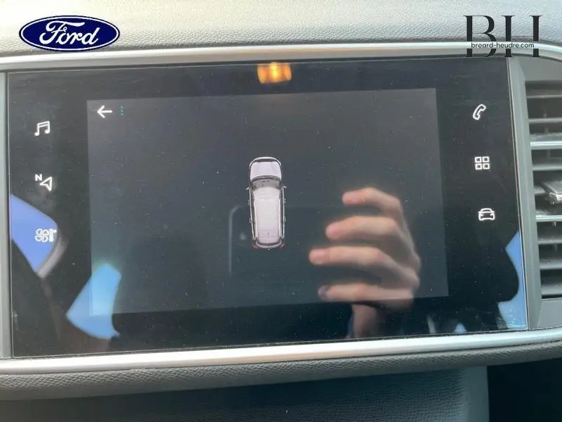 Écran tactile affichant la vue aérienne d'une Peugeot 308 SW gris Artense, avec reflet de main visible.