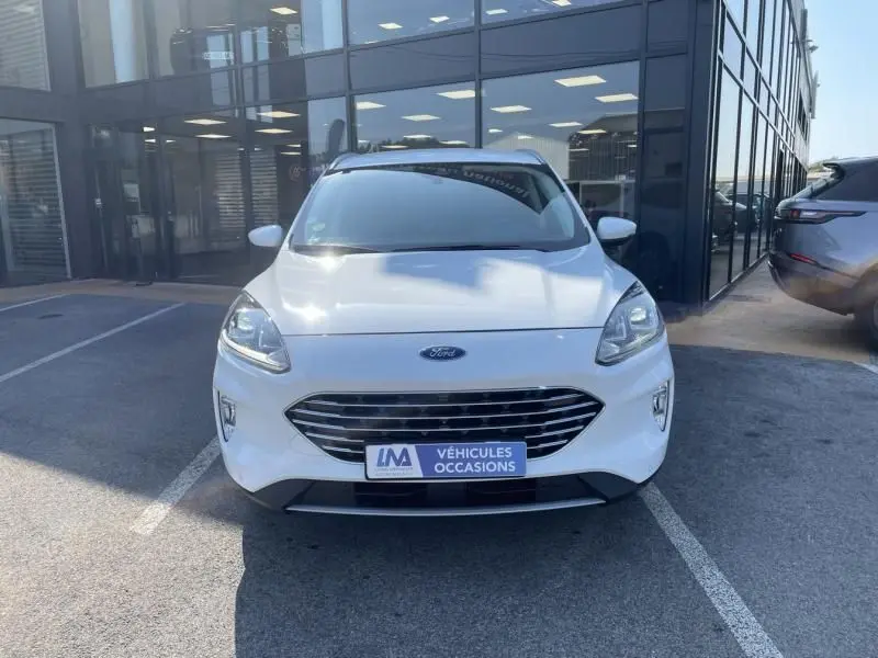 Vue frontale d'un Ford Kuga blanc 2020 avec calandre chromée et plaque "Véhicules Occasions" devant un bâtiment vitré.