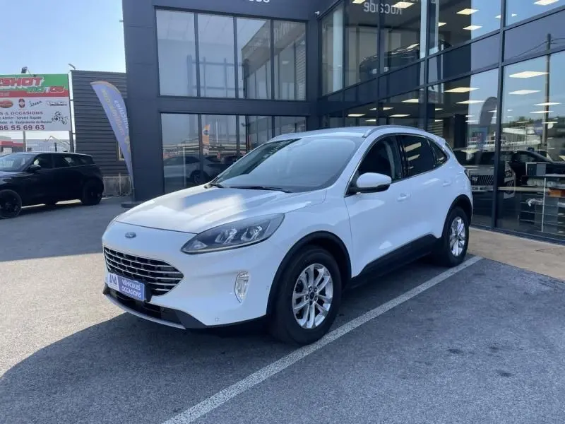 Ford Kuga blanc en 3/4 avant droit, version Titanium 2020, garé devant un bâtiment vitré en extérieur.