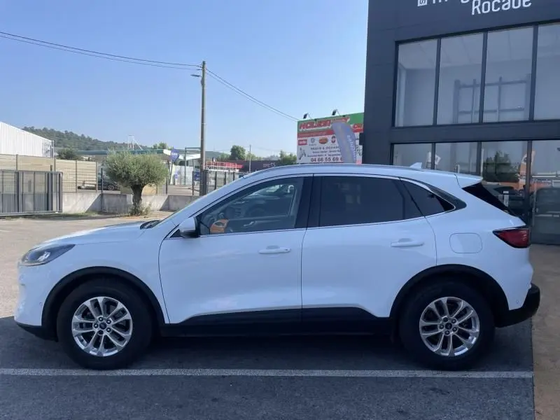 Vue latérale gauche d'un Ford Kuga blanc 2020 avec jantes alliage et vitres teintées, stationné devant un bâtiment moderne.