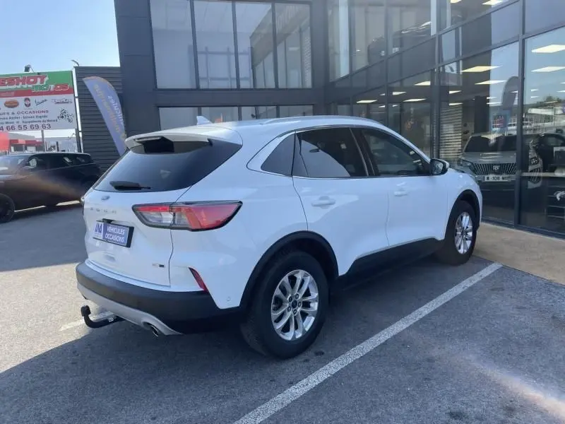 Vue 3/4 arrière droite d'un Ford Kuga blanc 2020 avec attelage, garé devant un bâtiment vitré.