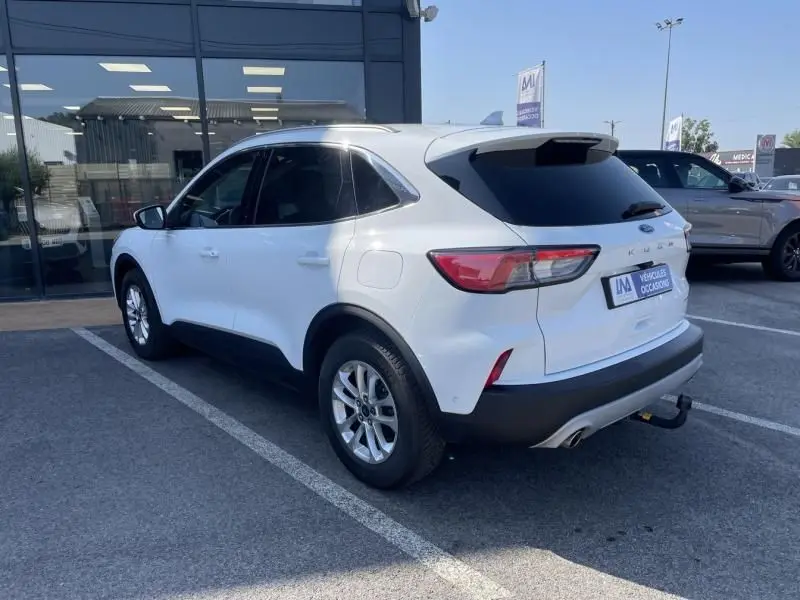 Vue 3/4 arrière droite d'un Ford Kuga blanc 2020 avec attelage et vitres arrière teintées, stationné devant un bâtiment.