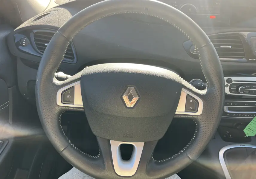 Vue rapprochée du volant en cuir noir avec logo Renault, tableau de bord et console centrale d'un Scenic III noir 2012.