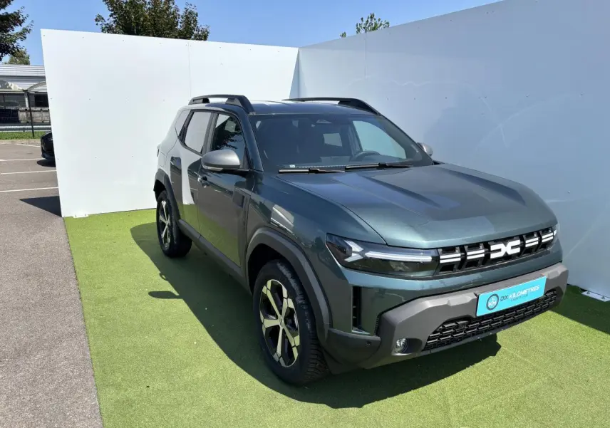 Dacia Duster III vert cèdre métal en 3/4 avant droit avec jantes alliage et barres de toit noires.