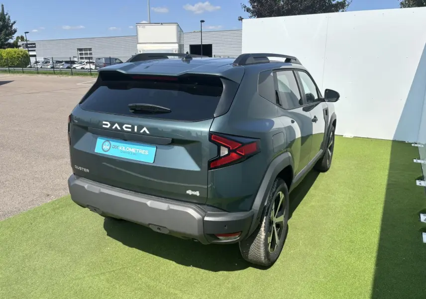 Vue 3/4 arrière droite du Dacia Duster 2025 vert cèdre métallisé avec jantes alliage et barres de toit.