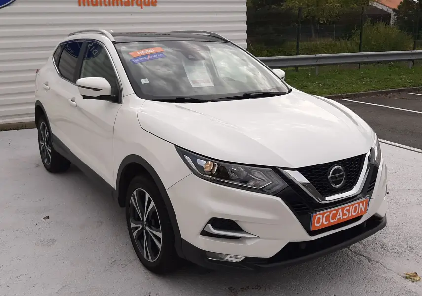 Nissan Qashqai blanc en 3/4 avant droit, avec plaques "OCCASION" et phares allumés sur parking extérieur.