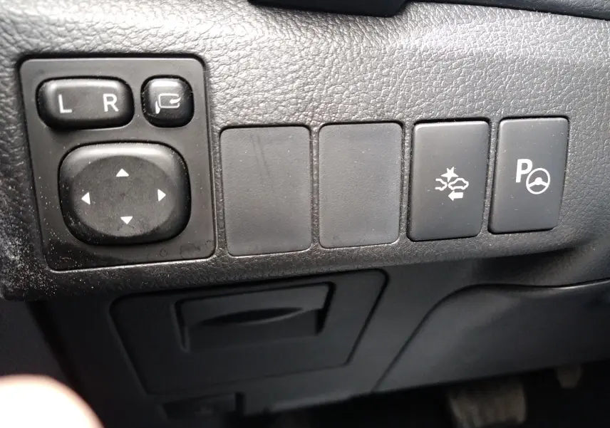 Gros plan sur les commandes de réglage des rétroviseurs et assistance au stationnement dans une Toyota Auris II hybride blanche de 2016.