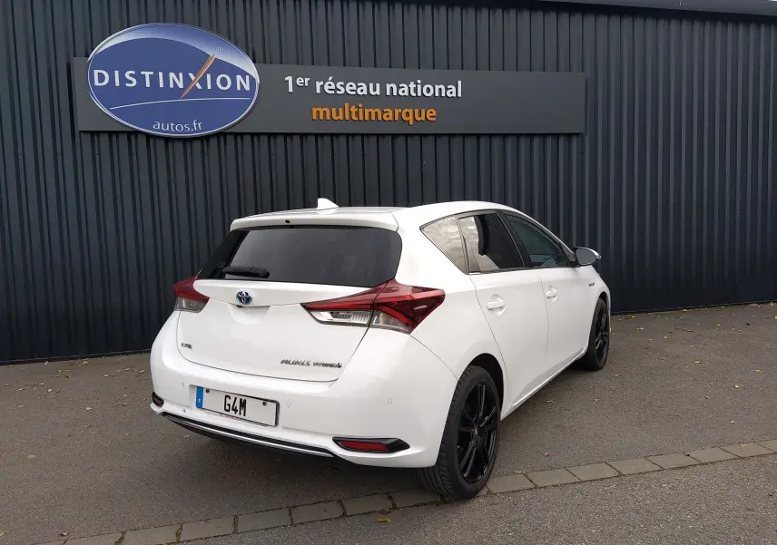 Toyota Auris hybride blanche vue 3/4 arrière droit, avec jantes noires et vitres teintées, stationnée devant un mur sombre.