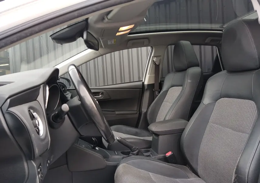 Vue intérieure côté conducteur de la Toyota Auris blanche, sièges en cuir noir avec inserts tissu gris et toit ouvrant panoramique.