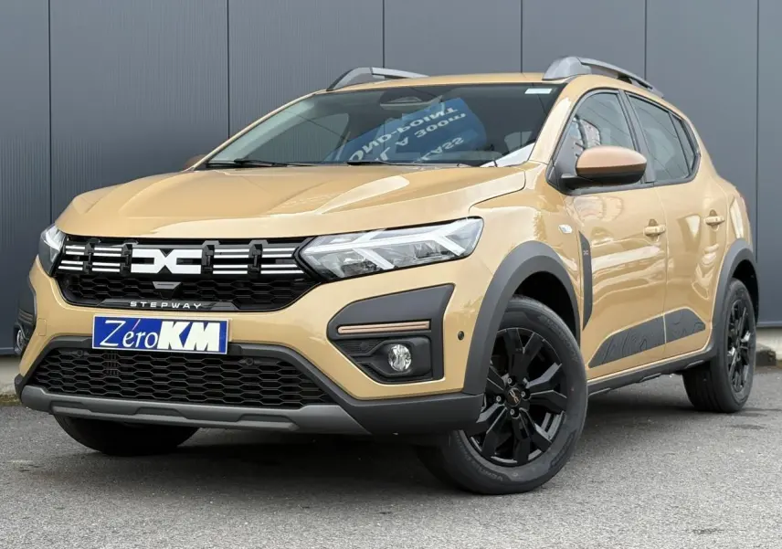 Vue 3/4 avant droit d'une Dacia Sandero Stepway 2025 beige Safari avec jantes noires et barres de toit gris Mégalithe.