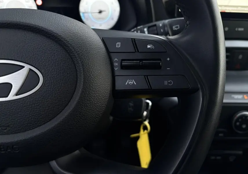 Gros plan sur le volant cuir noir de la Hyundai i20 2025 avec commandes au volant et compteur numérique flou en arrière-plan.