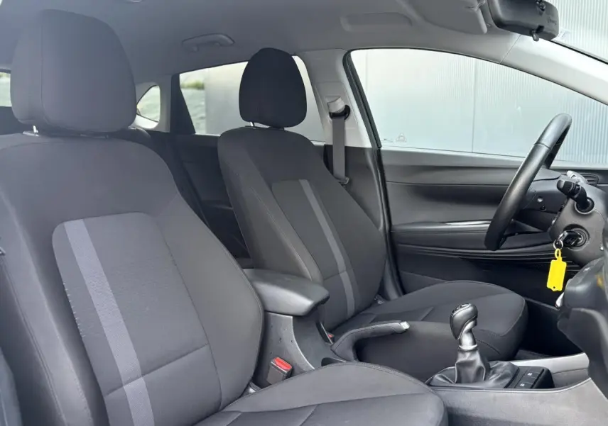 Vue intérieure côté droit de la Hyundai i20 2025, sièges tissu noirs avec accoudoir central et levier de vitesses manuel.