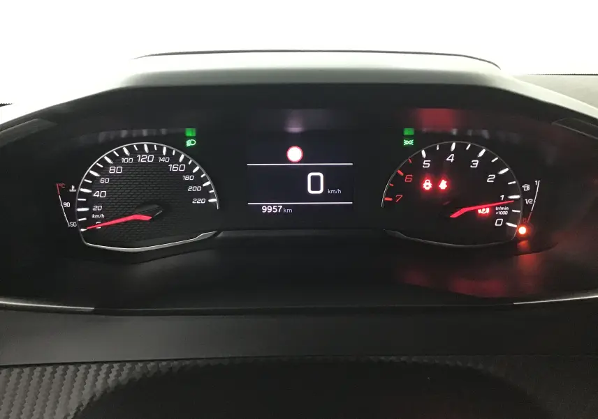 Tableau de bord numérique de la Peugeot 208 noire, affichant 0 km/h et 9957 km au compteur, avec voyants allumés.