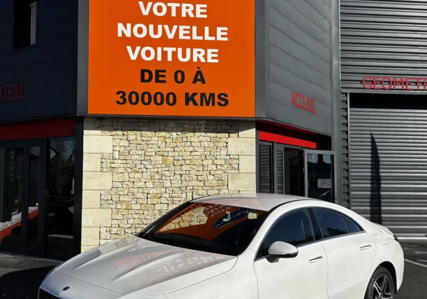 Mercedes CLA 250 e AMG Line blanc vue 3/4 avant droit, stationnée devant un bâtiment avec panneau orange Distinxion.