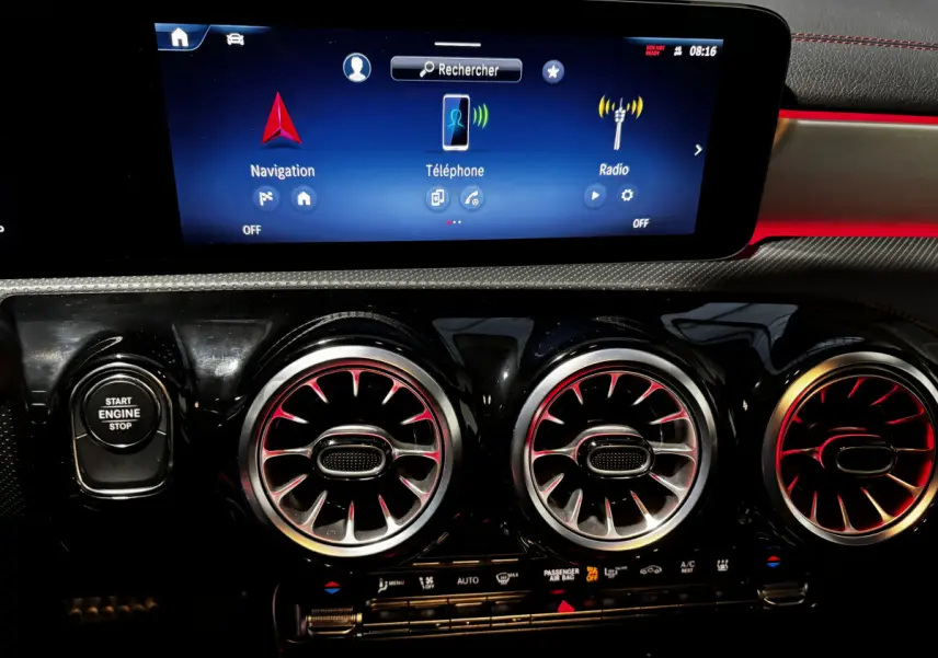 Vue rapprochée du tableau de bord noir avec écran tactile et aérateurs ronds à éclairage rouge d'une Mercedes CLA 250 e AMG Line blanche.