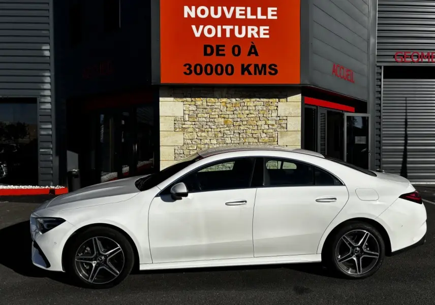Vue de profil côté gauche d'une Mercedes CLA 250 e AMG Line blanche stationnée devant un bâtiment avec panneau publicitaire rouge.