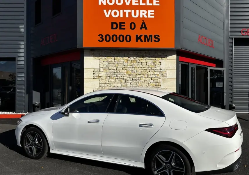 Vue de profil côté gauche d'une Mercedes CLA 250 e AMG Line blanche stationnée devant un bâtiment avec panneau orange.