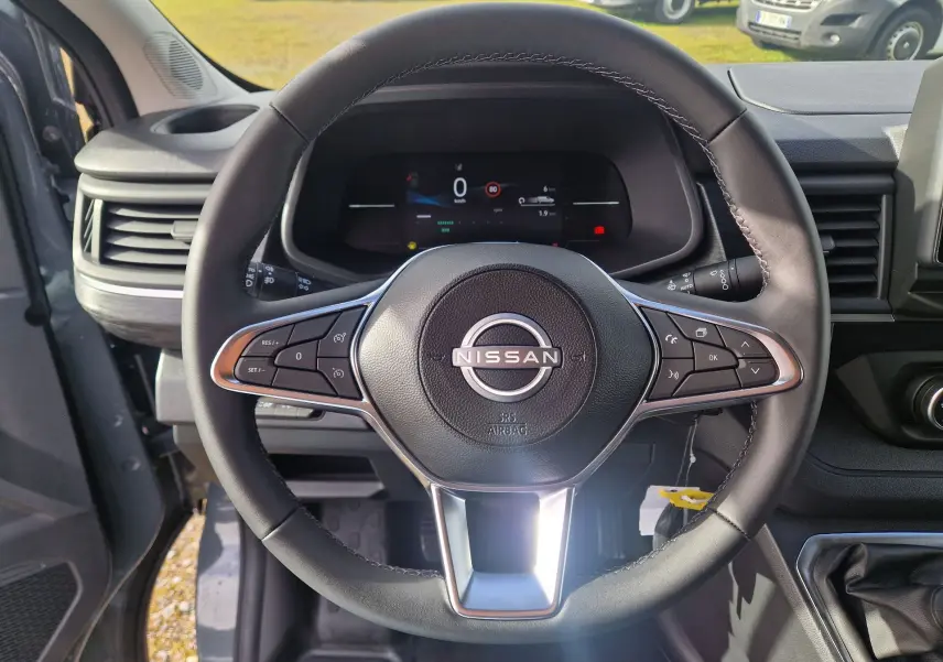 Vue intérieure centrée sur le volant noir du Nissan Primastar 2025 avec tableau de bord numérique et détails bois.