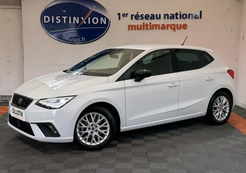Vue 3/4 avant droite d'une SEAT Ibiza blanche 2024 avec jantes alliage et phares LED allumés en intérieur showroom.