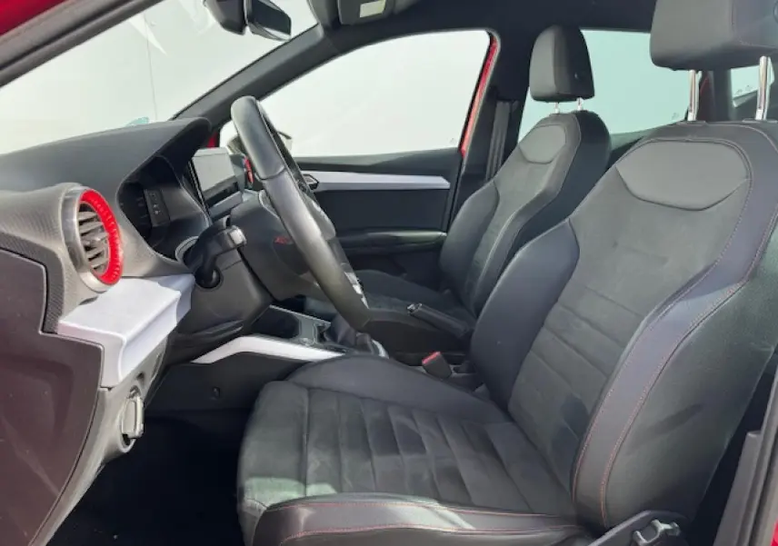 Vue intérieure côté conducteur du SEAT Arona 2024, sièges en suédine noire avec surpiqûres rouges et volant sport cuir.