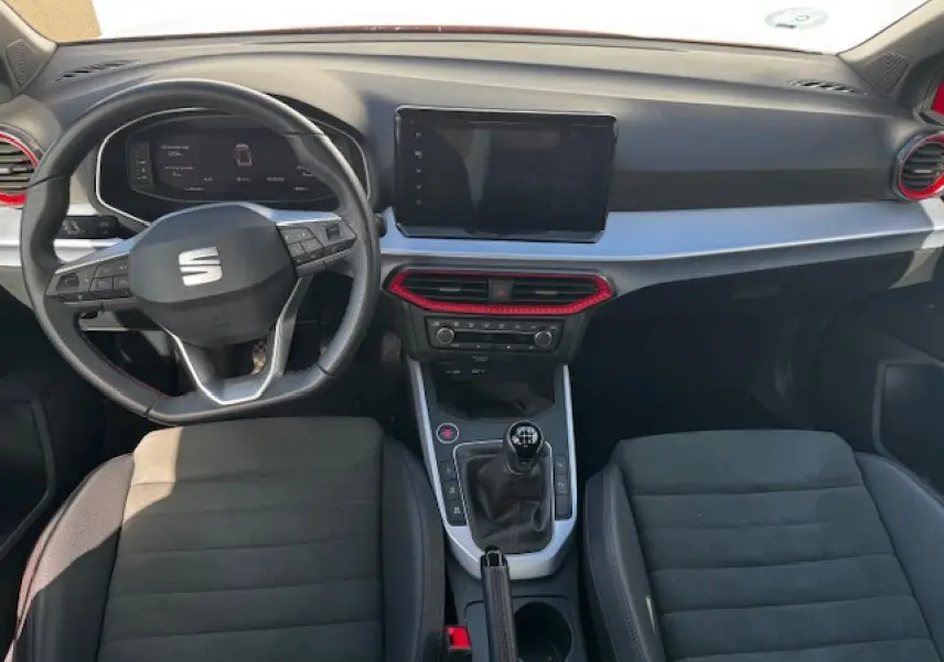Vue intérieure frontale du SEAT Arona 2024 avec volant cuir, console centrale manuelle et touches rouges décoratives.